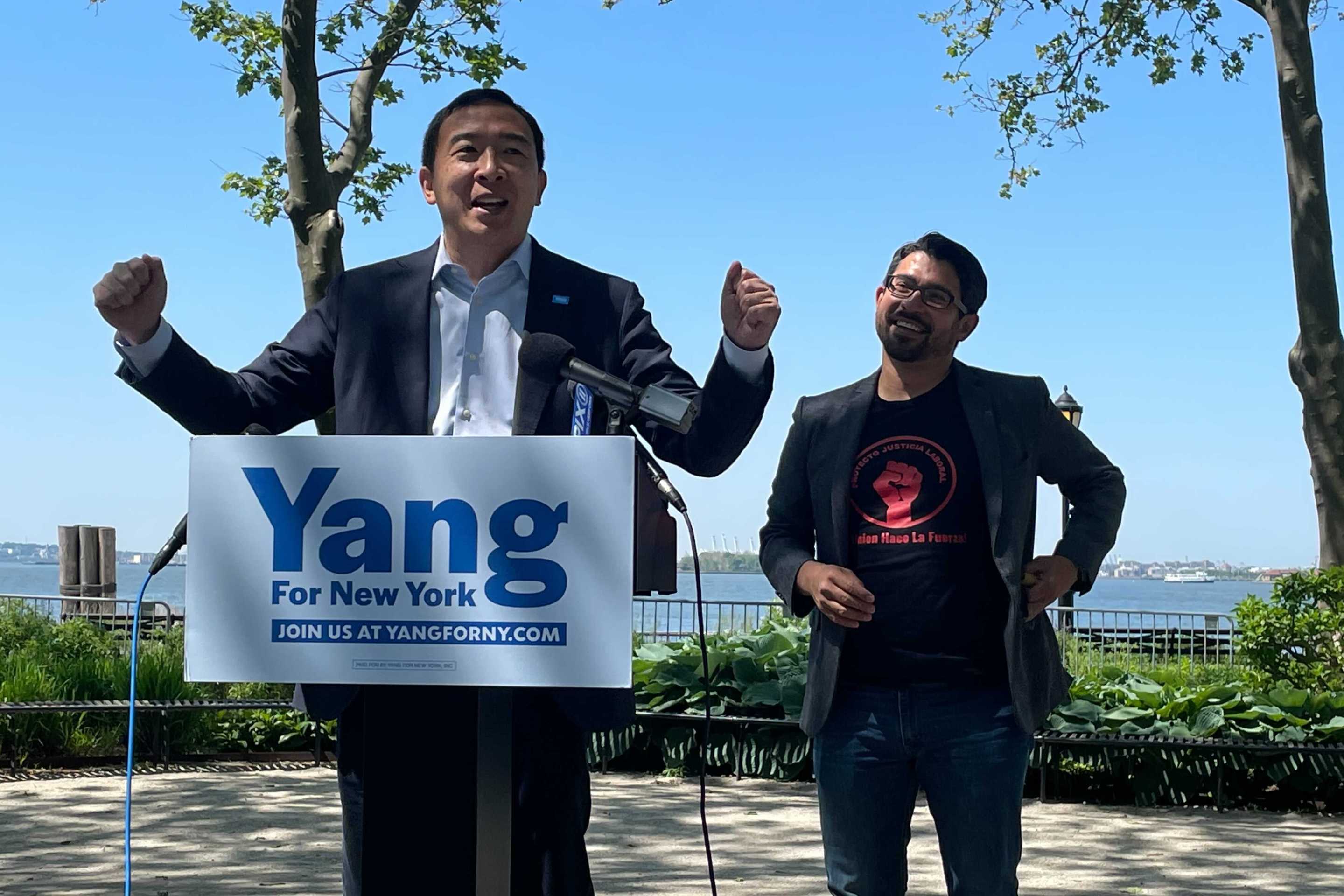 Andrew Yang with outstretched arms at a podium next to City Councilmember Carlos Menchaca in Battery Park with a view of the Statue of Liberty.