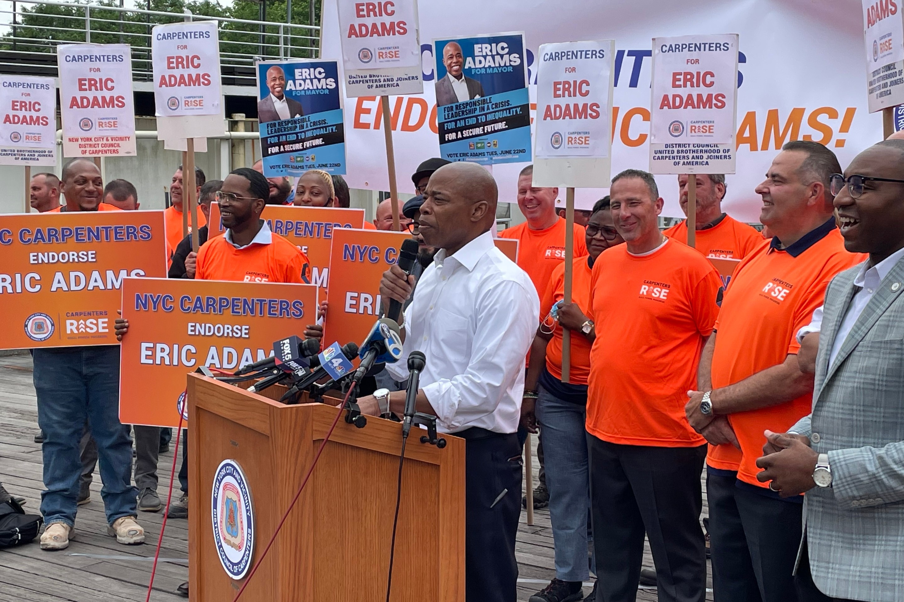 Eric Adams wears a white shirt and stands in front of a group of carpenters in Brooklyn Bridge Park as he receives their endorsement.