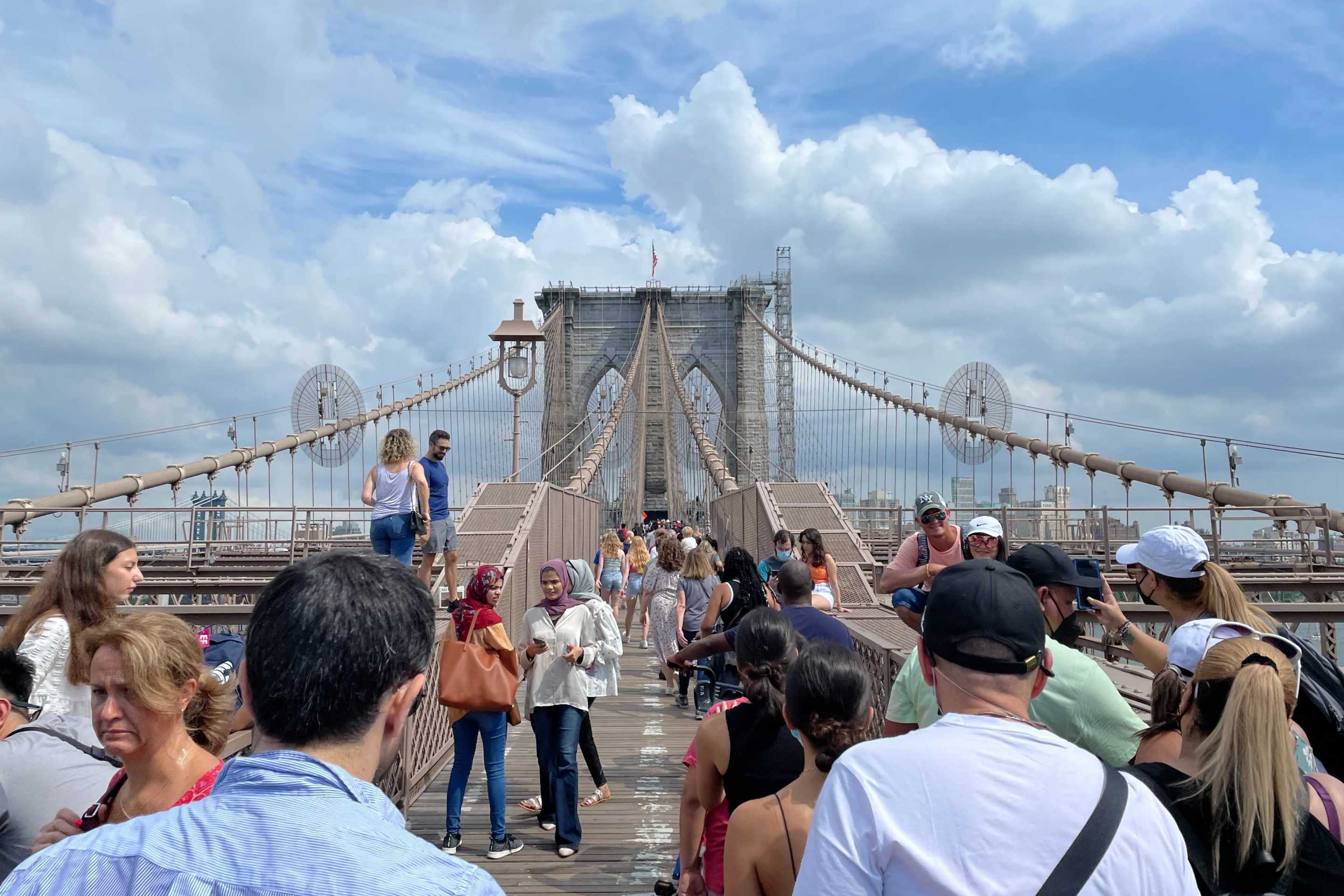 A large crowd of tourists stands on the Brooklyn Bridge walkway under sunny blue skies in July.