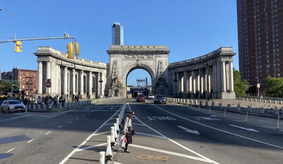 Wide daytime view of the Manhattan Bridge’s grand arched portal and flanking colonnade at Canal Street. Multiple traffic lanes and bike/parking separators lead toward the arch; a few pedestrians stand near the curb in the foreground and cars drive through the arch. Tall buildings and a clear blue sky are visible beyond and beside the structure.