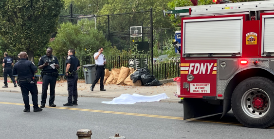 On a city street, several uniformed emergency responders gather near a body covered by a white sheet on the sidewalk. An FDNY fire truck is parked at the right edge of the frame. Trash bags and a fenced basketball court with trees are visible in the background.