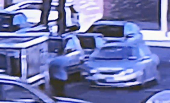 Grainy surveillance photo of a silver SUV on a sidewalk near parked vehicles and a street tree; a blurred person appears in front of the vehicle and another figure is nearby. Buildings and a storefront are visible in the background.